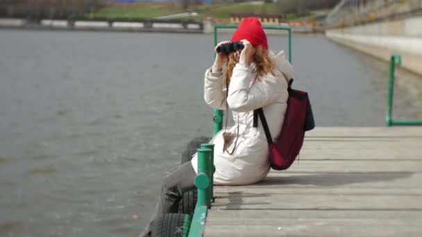 Una hermosa mujer con una chaqueta y un sombrero rojo está de pie en el muelle y mirando a través de binocular — Vídeos de Stock