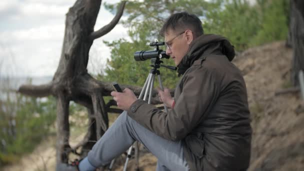 Alter Mann blickt durch ein Teleskop in einem Wald am Flussufer — Stockvideo