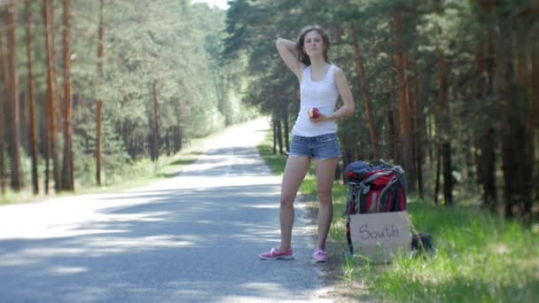 Giovane bella donna autostop in piedi sulla strada con uno zaino su un tavolo con un'iscrizione SUD — Video Stock