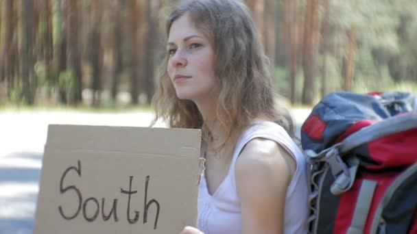 Молода красива жінка автостоп стоїть на дорозі з рюкзаком на столі з написом SOUTH — стокове відео