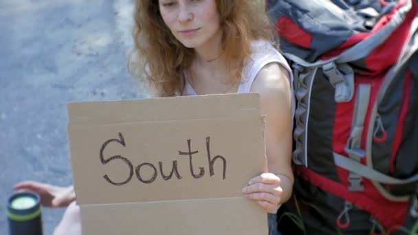 Young beautiful woman hitchhiking standing on the road with a backpack on a table with an inscription SOUTH — Stock Video