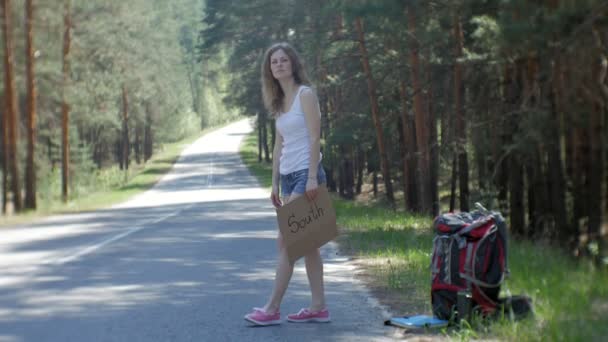 Fiatal gyönyörű nő autóstoppal áll az úton egy hátizsák, egy asztal, egy Felirat Dél — Stock videók