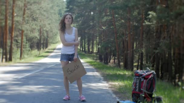 Mulher bonita jovem carona em pé na estrada com uma mochila em uma mesa com uma inscrição SOUTH — Vídeo de Stock