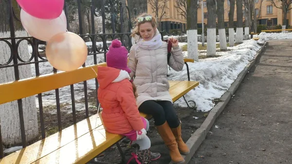 Una hermosa niña y su madre están sentadas en un banco del parque y comiendo un delicioso postre y bebiendo té de un termo a principios de primavera — Foto de Stock