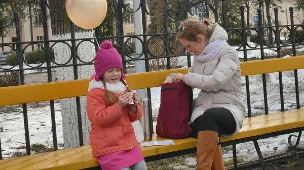 Красивая девушка и ее мама сидят на скамейке в парке и едят вкусный десерт и пьют чай из термоса ранней весной — стоковое фото