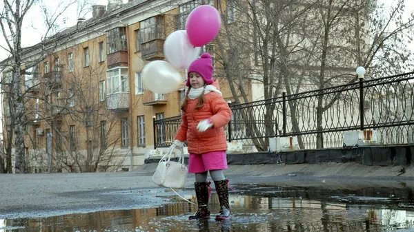 Pequena menina bonita pulando em poças após a chuva com balões coloridos — Fotografia de Stock