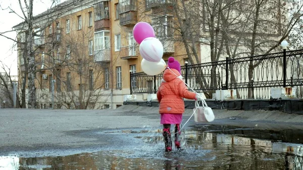Маленькая красивая девочка прыгает на лужах после дождя с красочными воздушными шарами — стоковое фото