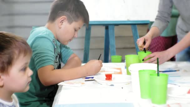 Діти хлопчики та дівчатка сидять разом за столом у класі та малюють. З ними їхній молодий і красивий вчитель . — стокове відео