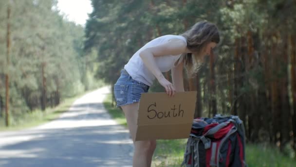 Młoda kobieta piękne autostopem stojący na drodze z plecakiem na stół z napisem południe — Wideo stockowe