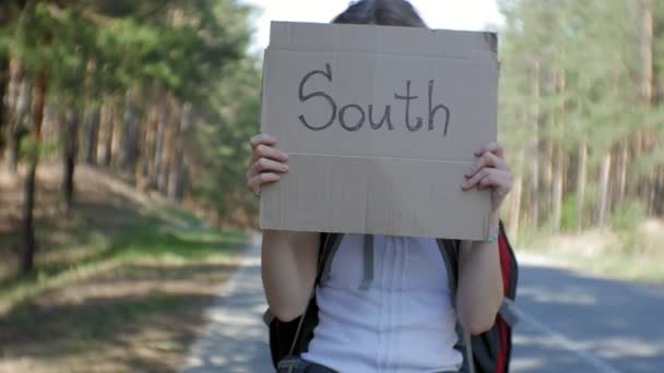 Young beautiful woman hitchhiking standing on the road with a backpack on a table with an inscription SOUTH — Stock Video