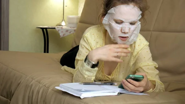 Junge Frau macht Gesichtsmaske mit Reinigungsmaske, klickt zu Hause mit Smartphone auf Couch — Stockfoto