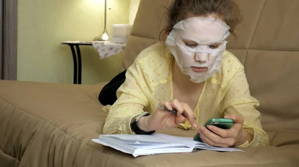 Jonge vrouw doet gezichtsmasker met reinigende masker, masker klikken op Bank met smartphone thuis — Stockfoto