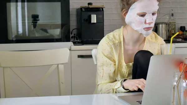 Jonge vrouw doet gezichtsmasker met zuivering masker, masker werken achter een laptop thuis — Stockfoto