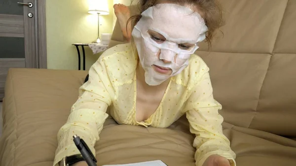 Wanita muda bertopeng wajah dengan topeng pembersih, klik di sofa dengan smartphone di rumah — Stok Foto