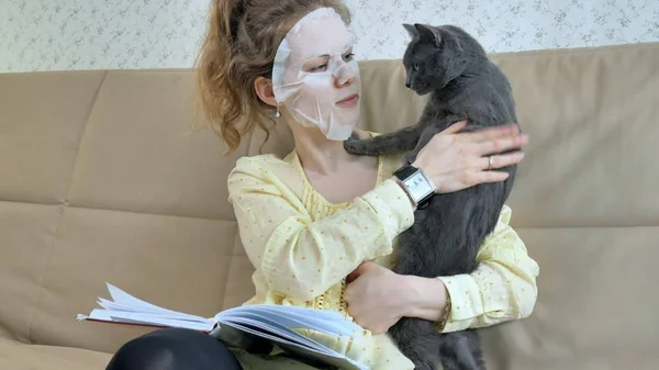 Young woman doing facial mask mask with cleansing mask on face in kitchen at home cat