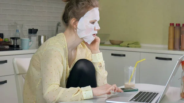 自宅のノート パソコンの背後に働くマスク、クレンジングと化粧水マスクを行う若い女性 — ストック写真