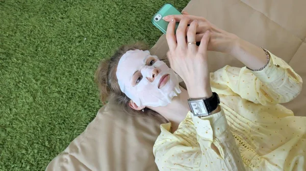 Mujer joven haciendo máscara facial con máscara de limpieza, clics en el sofá con teléfono inteligente en casa — Foto de Stock