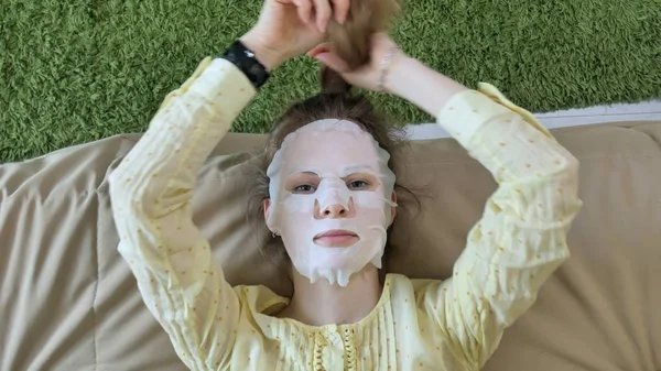 Mujer joven haciendo máscara facial con máscara de limpieza, clics en el sofá con teléfono inteligente en casa —  Fotos de Stock