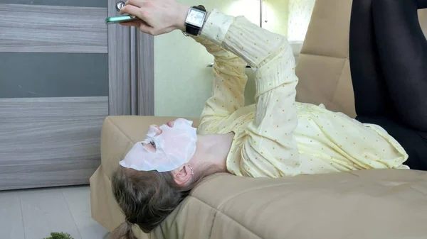 Jovem mulher fazendo máscara facial com máscara de limpeza, cliques no sofá com smartphone em casa — Fotografia de Stock