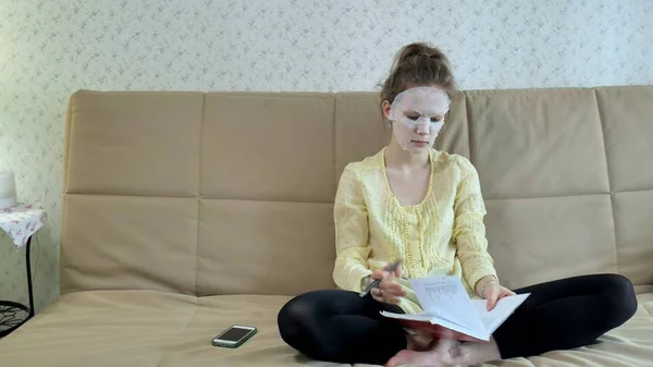 Ung kvinna gör ansiktsmask mask med rengörande mask, klickar på soffan med smartphone hemma — Stockfoto
