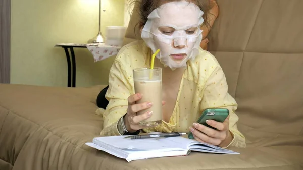 Jonge vrouw doet gezichtsmasker met reinigende masker, masker klikken op Bank met smartphone thuis — Stockfoto