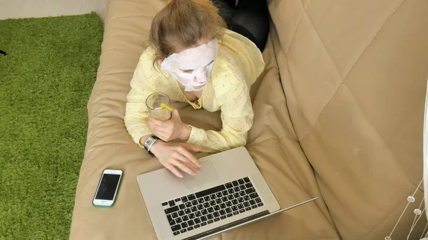 Giovane donna che fa maschera facciale con maschera detergente, lavorando dietro il computer portatile a casa — Foto Stock