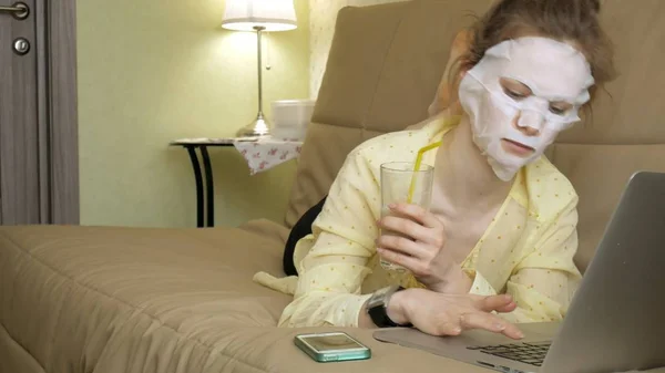 Giovane donna che fa maschera facciale con maschera detergente, lavorando dietro il computer portatile a casa — Foto Stock