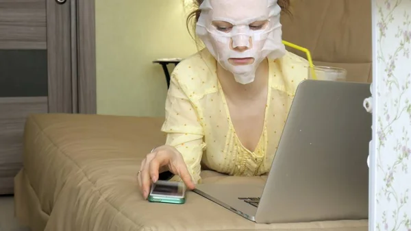 Jonge vrouw doet gezichtsmasker met zuivering masker, masker werken achter een laptop thuis — Stockfoto