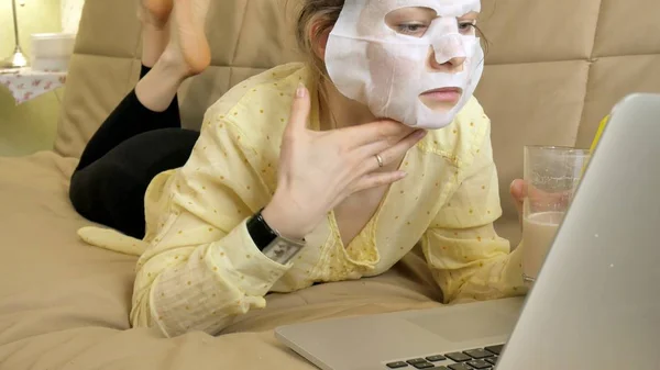 Молода жінка робить маску для обличчя з маскою для очищення, працюючи позаду ноутбука вдома — стокове фото
