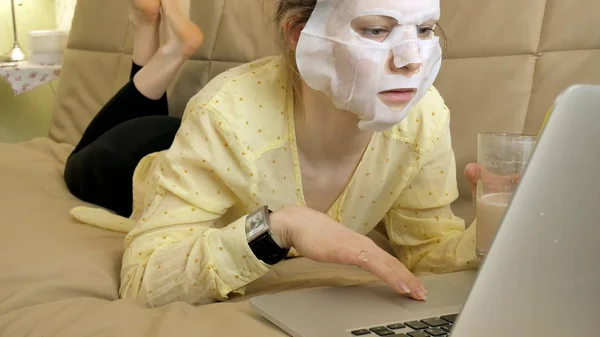 Nuori nainen tekee kasvonaamio puhdistava naamio, takana kannettavan tietokoneen kotona — kuvapankkivalokuva