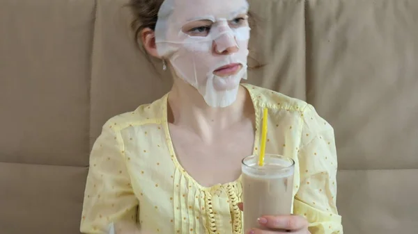 Mujer joven haciendo máscara facial con máscara de limpieza, clics en el sofá con teléfono inteligente en casa — Foto de Stock