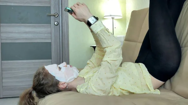 Jovem mulher fazendo máscara facial com máscara de limpeza, cliques no sofá com smartphone em casa — Fotografia de Stock