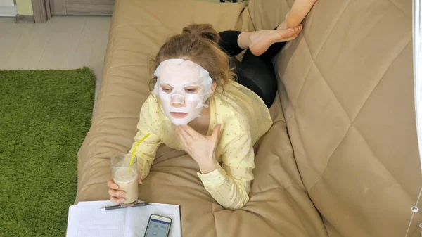 Молода жінка робить маску для обличчя з маскою для очищення, клацає на дивані зі смартфоном вдома — стокове фото
