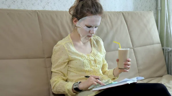 Wanita muda bertopeng wajah dengan topeng pembersih, klik di sofa dengan smartphone di rumah — Stok Foto