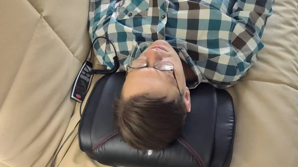 Homem deita-se no sofá em casa, sob a cabeça de um massageador, estado relaxado — Fotografia de Stock