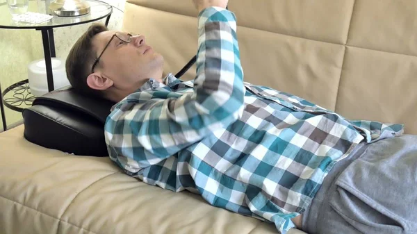 Mies makaa sohvalla kotona, hierontalaitteen pään alla, rento tila — kuvapankkivalokuva