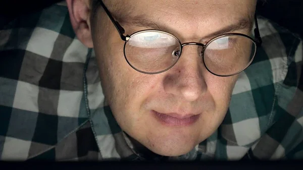 Um homem de óculos trabalha até tarde da noite. Ele parece preocupado, vê na tela do computador na frente dele, um close-up — Fotografia de Stock