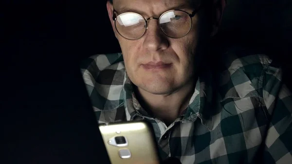 Um homem de óculos trabalha até tarde da noite. Ele parece preocupado, vê na tela do computador na frente dele, um close-up — Fotografia de Stock