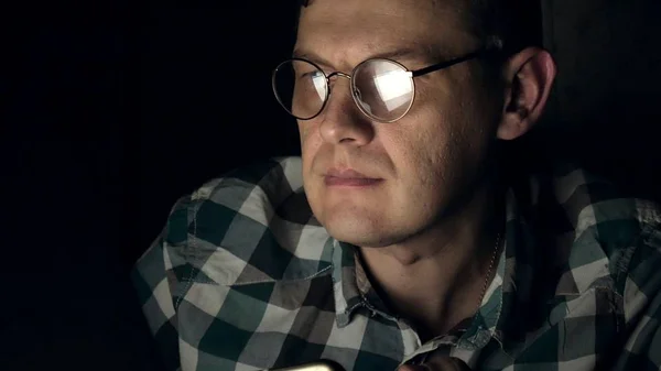 Un hombre con gafas trabaja hasta tarde por la noche. Se ve preocupado, ve en la pantalla de la computadora frente a él, un primer plano —  Fotos de Stock