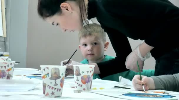 Niños niños sentados juntos alrededor de la mesa en el aula y dibujando. Con ellos está su joven y hermosa maestra . — Vídeos de Stock