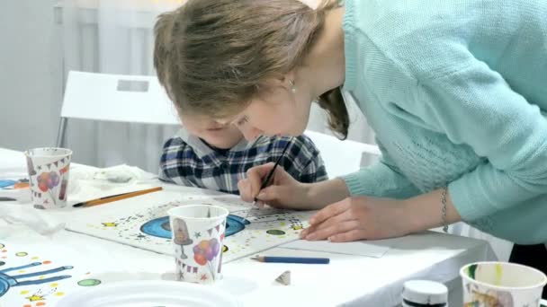 Bambini ragazzi seduti insieme intorno al tavolo in classe e disegno. Con loro c'è la loro giovane e bella maestra . — Video Stock