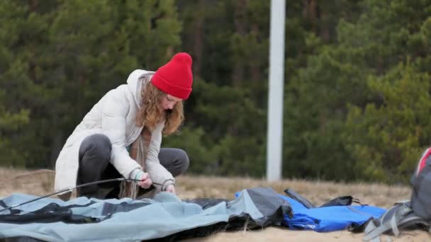 Mulher turística jovem atraente em um chapéu vermelho recolhe uma barraca turística perto da floresta na costa — Vídeo de Stock