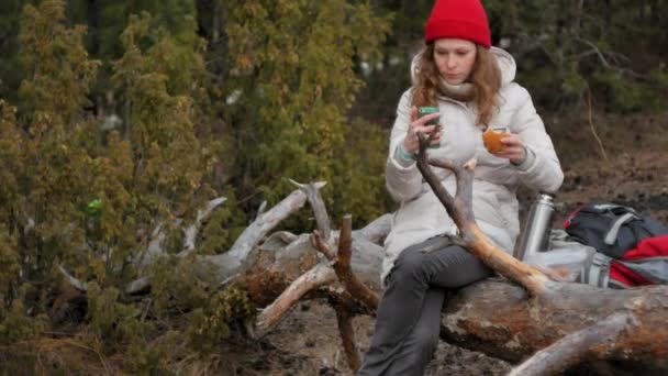 Приваблива молода жінка в червоному капелюсі проходить через ліс ранньою весною з великим туристичним рюкзаком, п'є чай з термоса — стокове відео