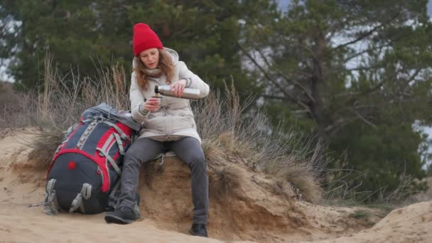 Приваблива молода жінка в червоному капелюсі проходить через ліс ранньою весною з великим туристичним рюкзаком, п'є чай з термоса — стокове відео