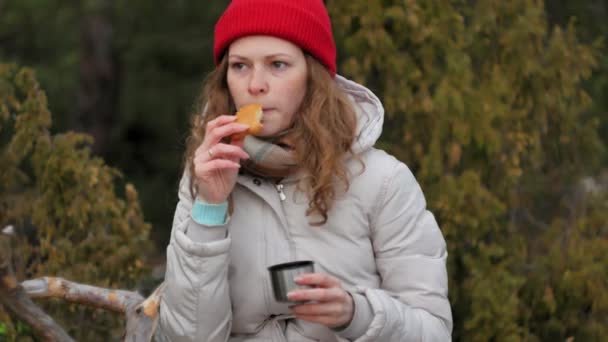 An attractive young woman in a red hat walks through the forest in early spring with a large tourist backpack, drinks tea from a thermos — Stock Video