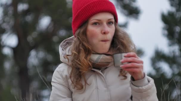 En attraktiv ung kvinna i en röd hatt promenader genom skogen tidigt på våren med en stor turist ryggsäck, drycker te från en termos — Stockvideo