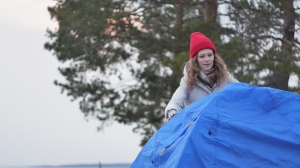 Attraente giovane donna turistica con un cappello rosso raccoglie una tenda turistica vicino alla foresta sulla costa — Video Stock