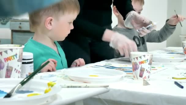 Niños niños sentados juntos alrededor de la mesa en el aula y dibujando. Con ellos está su joven y hermosa maestra . — Vídeos de Stock