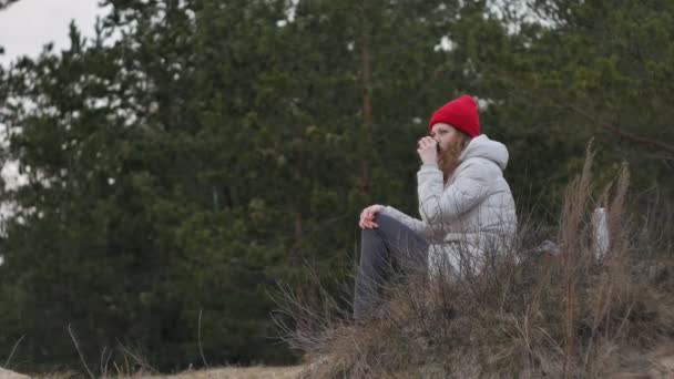 Une jolie jeune femme au chapeau rouge se promène dans la forêt au début du printemps avec un grand sac à dos touristique, boit du thé dans un thermos — Video