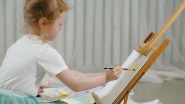 Hermosa chica pelirroja se sienta en el suelo en el estudio delante del caballete y dibuja un pincel y pinturas de colores — Vídeos de Stock